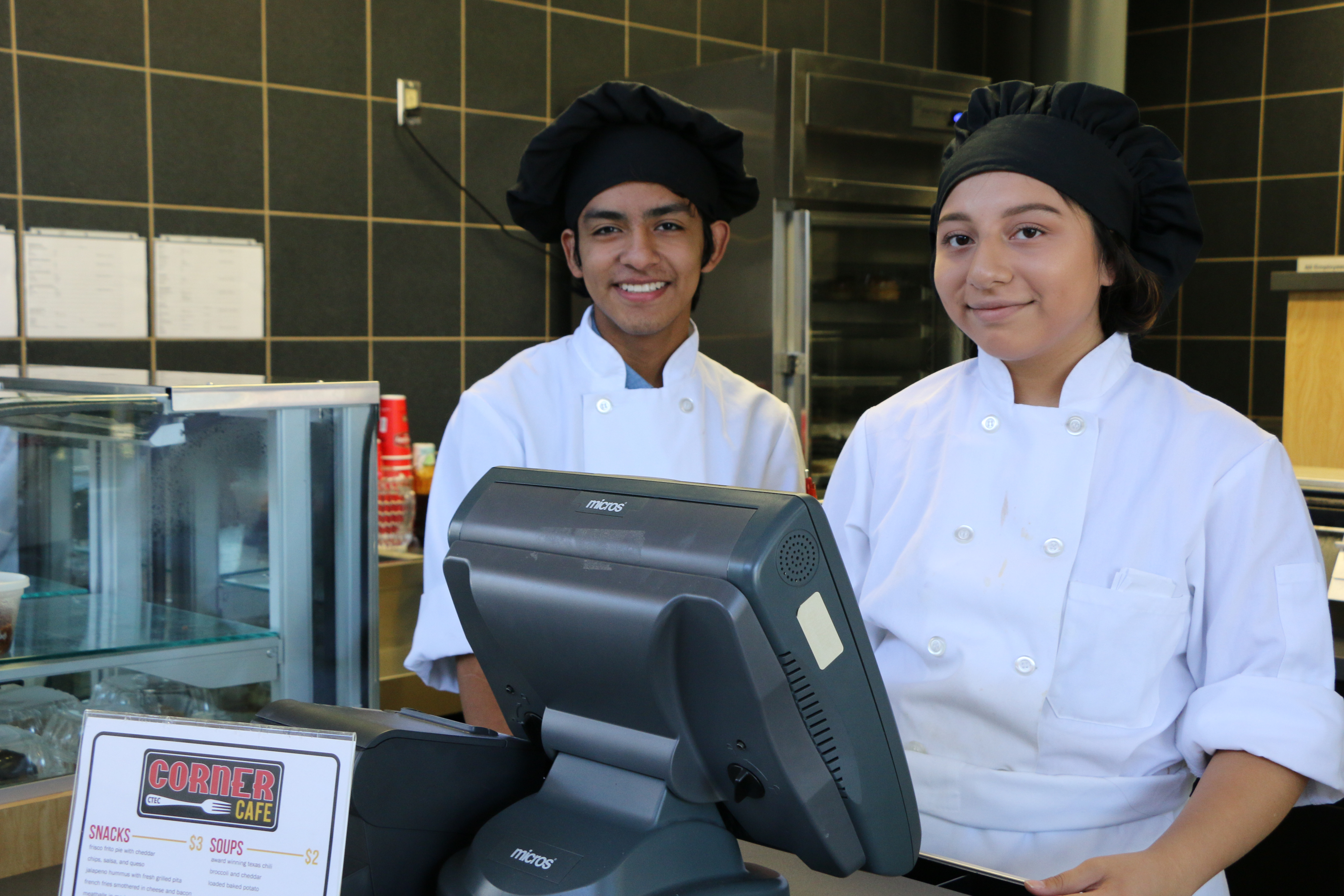 Two culinary students are stationed at the cash register of an eating establishment. They wear white chefs coats and black chefs hats.