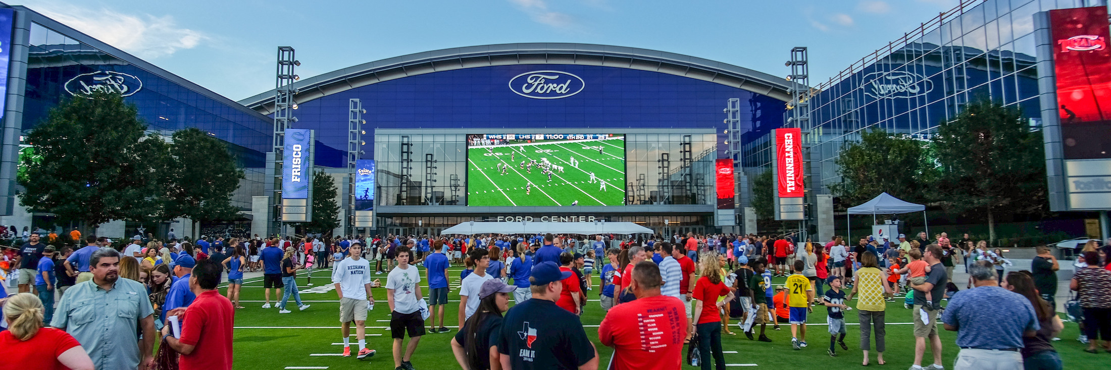People gather on Tostito's Plaza in front of Ford Center at The Star. There is a football game shown on the large screen on the building.