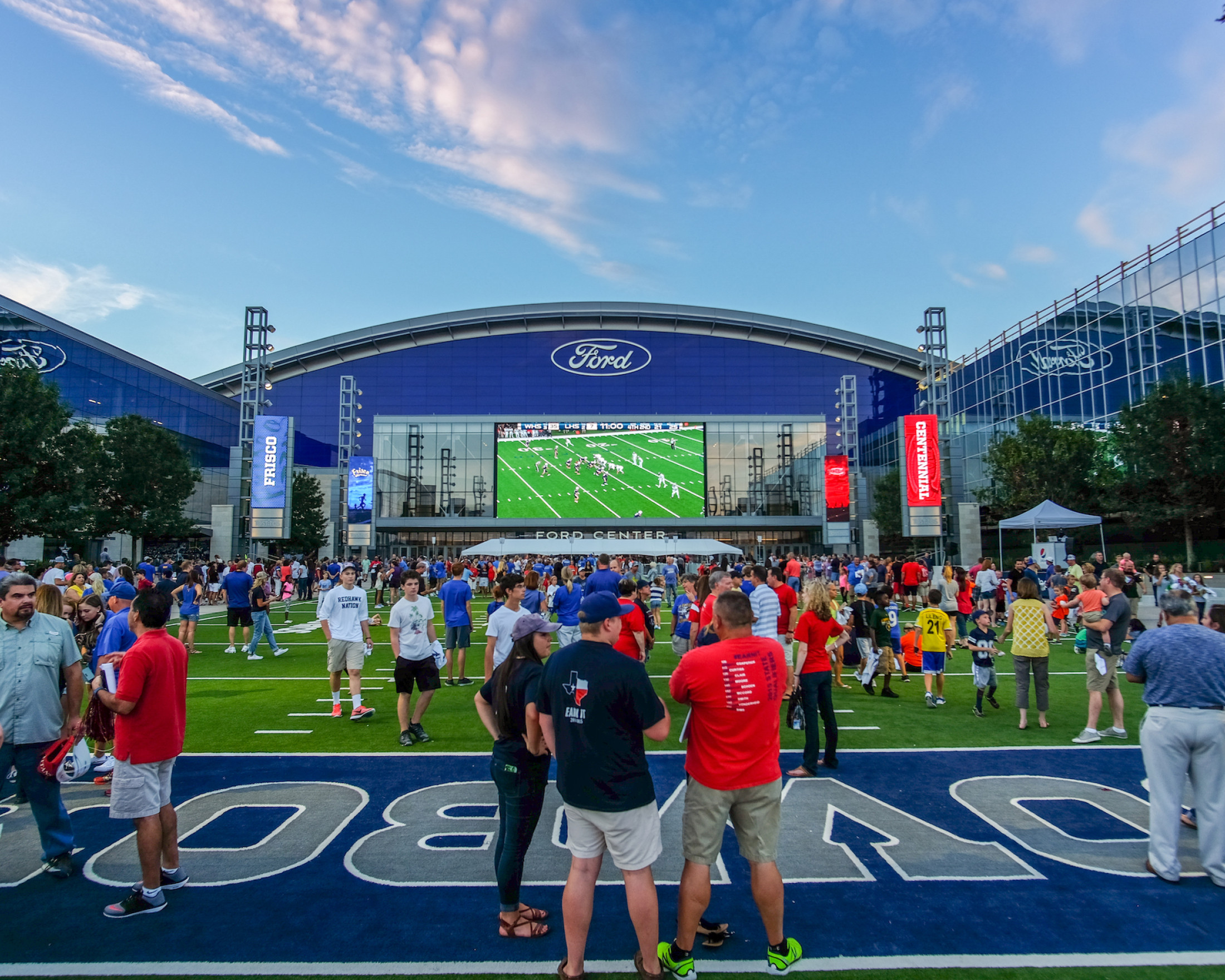 People gather on Tostito's Plaza in front of Ford Center at The Star. There is a football game shown on the large screen on the building.