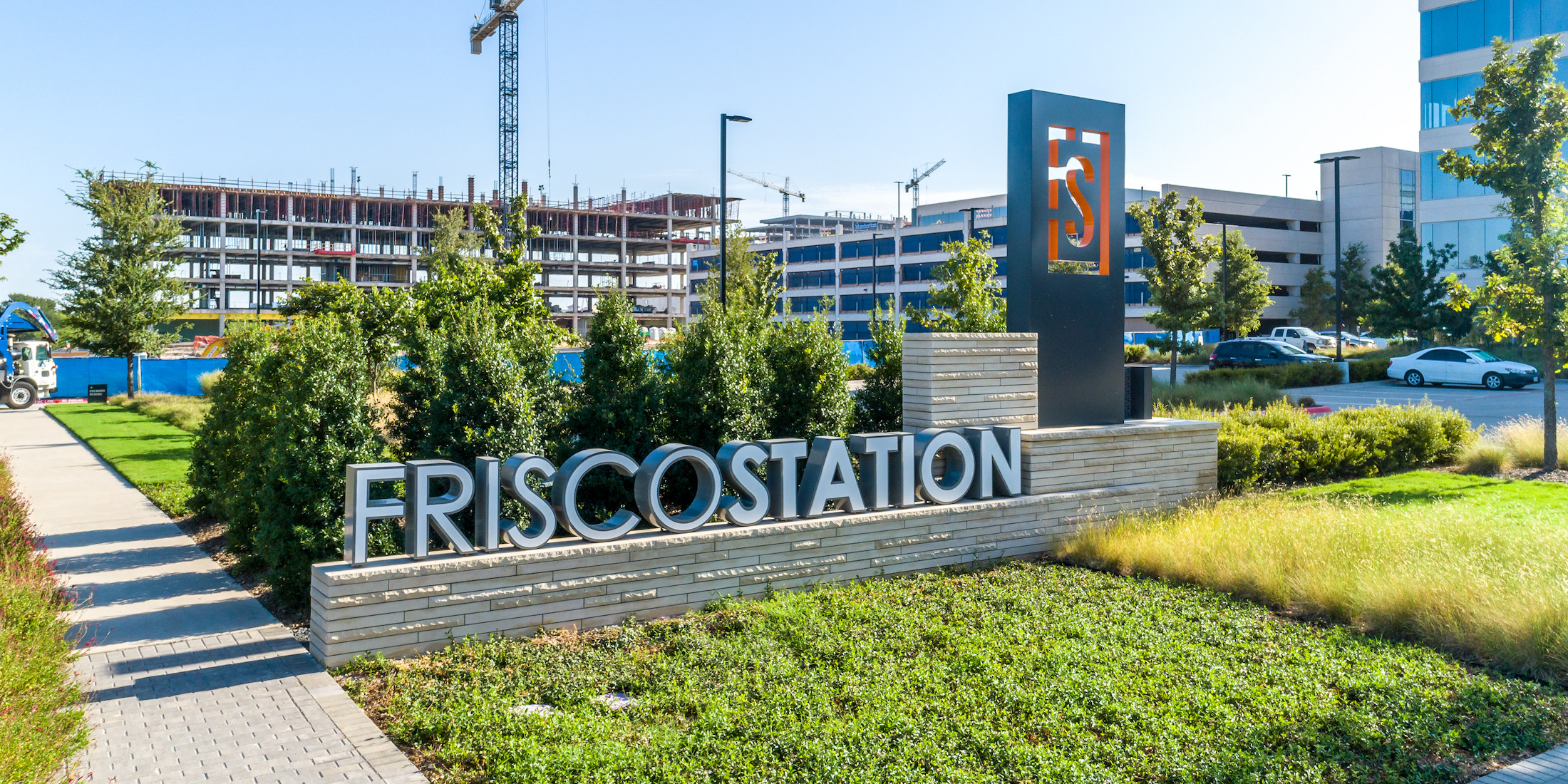 Photo of the Frisco Station sign. There are office buildings under construction in the background.