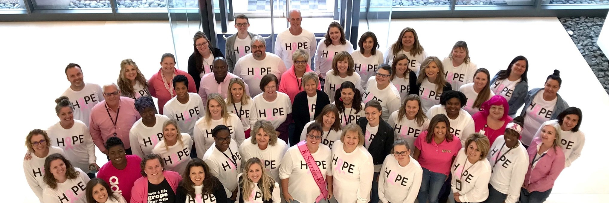 A large group of people are gathered in the lobby of a building looking up and smiling at the camera which is on the level above them.