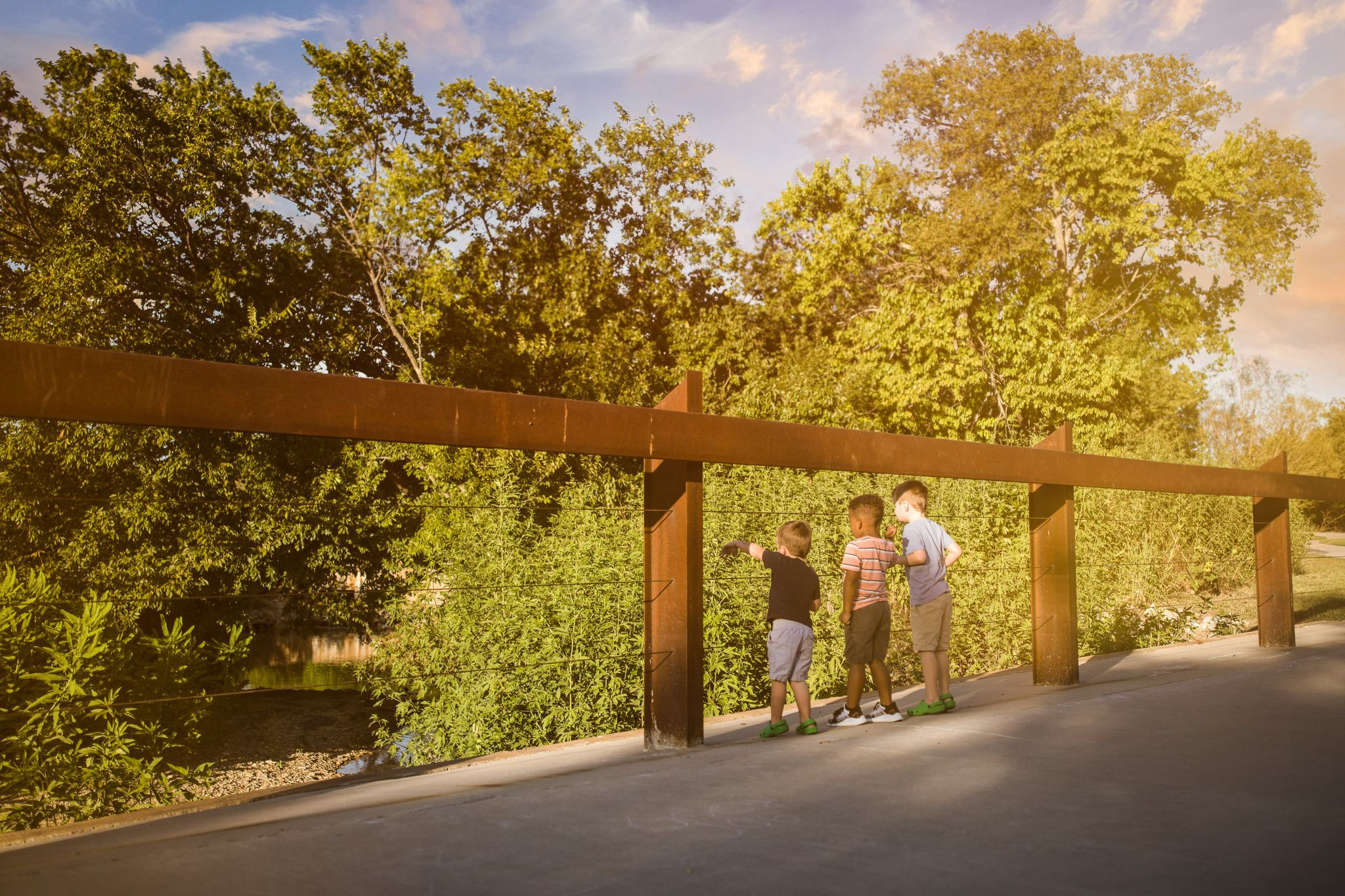 Three young boys stand together on a bridge looking at water down below.