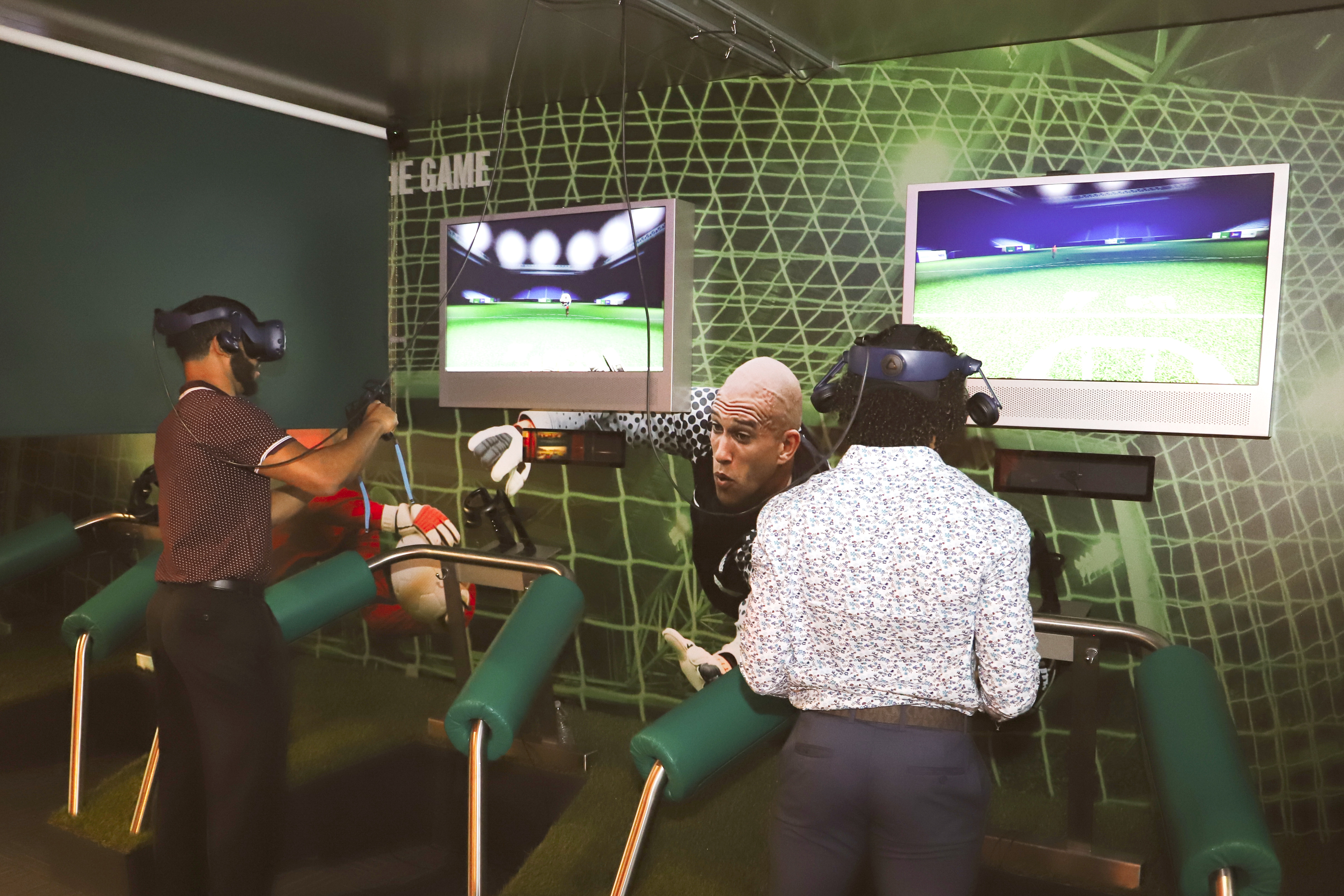 Two men play a virtual reality game at the National Soccer Hall of Fame. They both wear headsets that go over their eyes and they hold remotes in each hand.