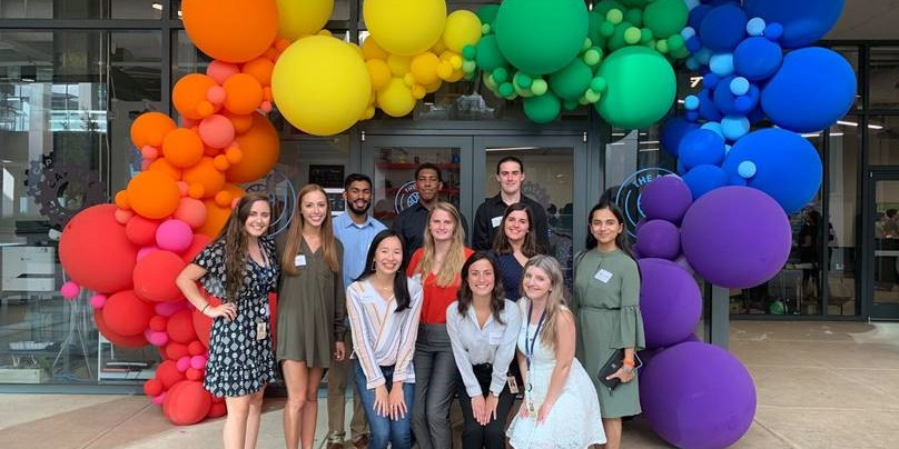 A group of 11 people pose outdoors for a picture in front of a building with a rainbow balloon arch.