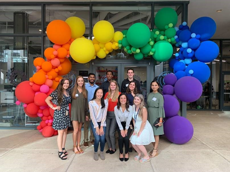 A group of 11 people pose outdoors for a picture in front of a building with a rainbow balloon arch.