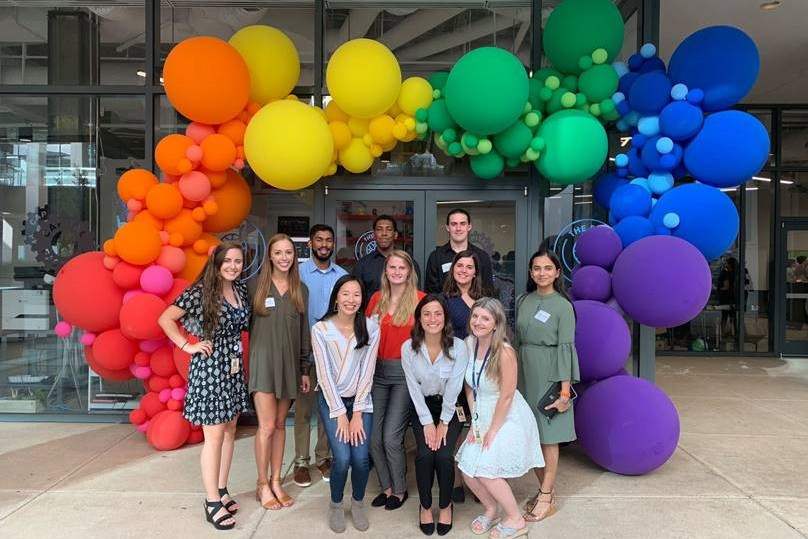 A group of 11 people pose outdoors for a picture in front of a building with a rainbow balloon arch.
