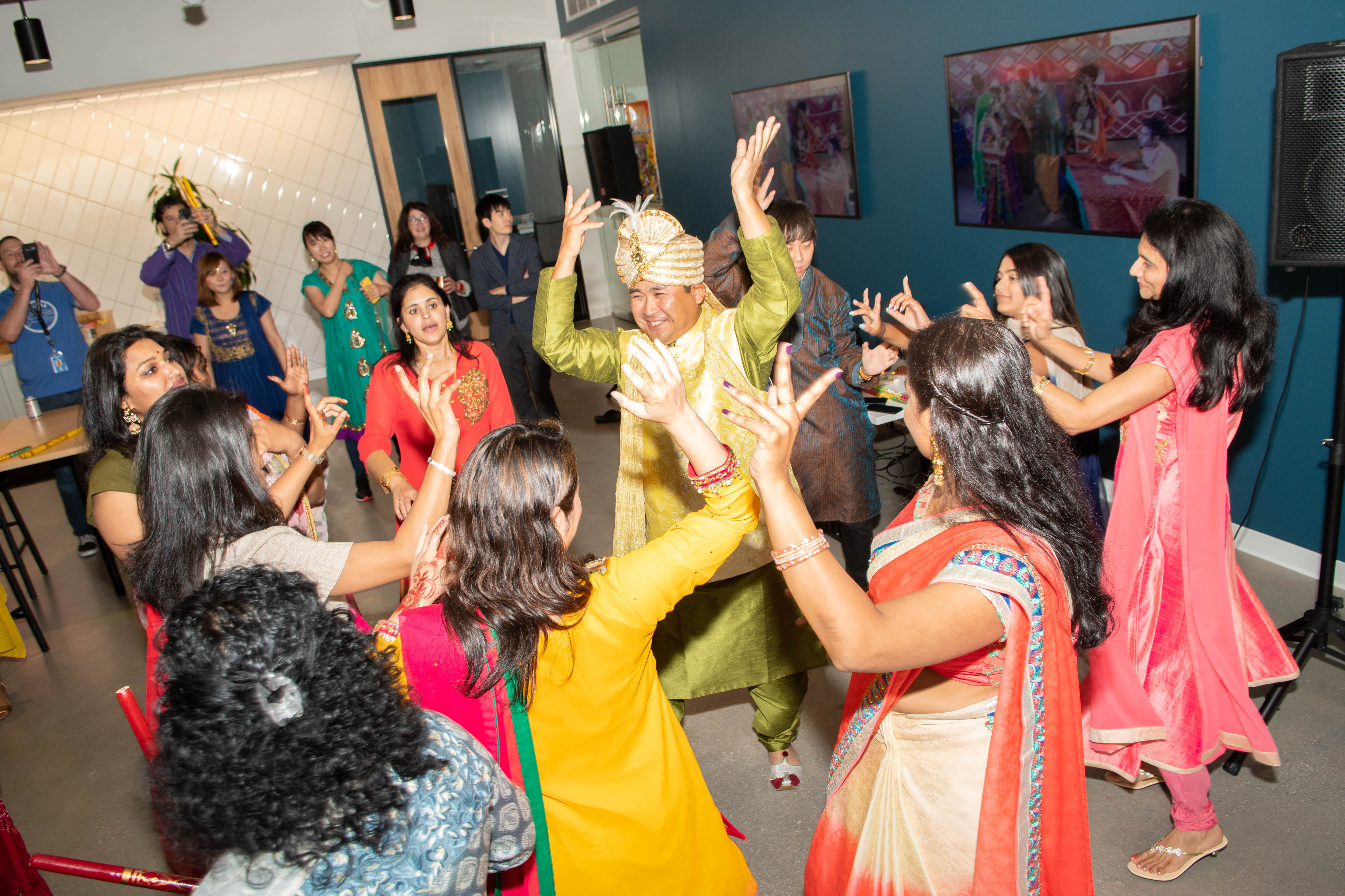 A group of people dancing indoors. They wear colorful clothing. This is part of a Diwali celebration.
