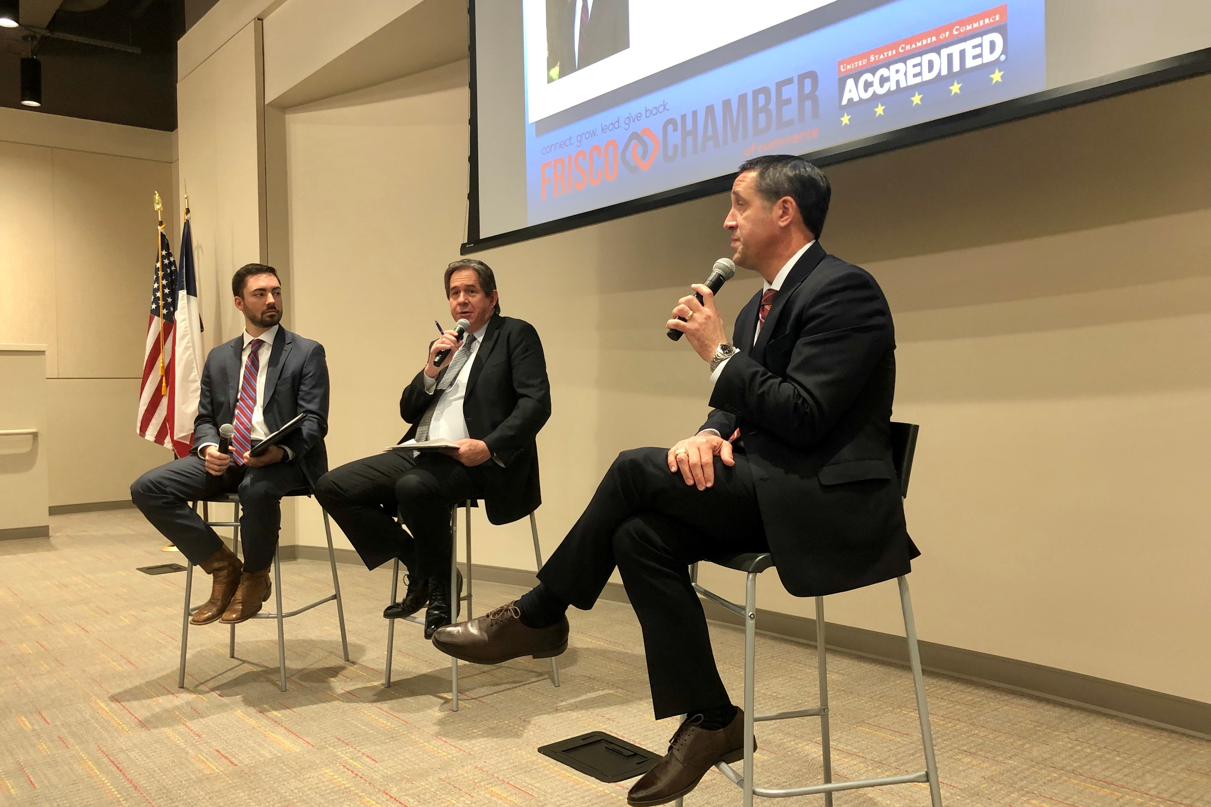 Three representatives from the Frisco Chamber of Commerce sit in chairs at the front of an event. They all hold microphones.