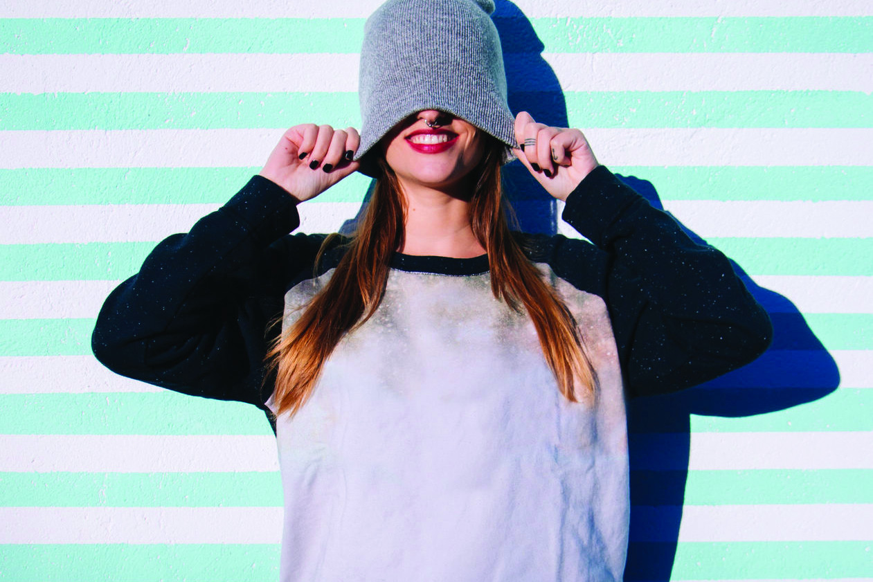 A girl against a striped wall pulls her beanie over her head and smiles.