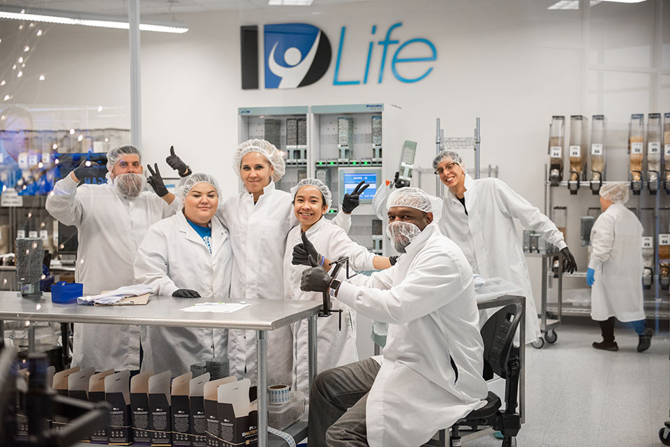 Six lab workers gather for a photo in the lab at ID Life. They wear lab coats and hair nets.
