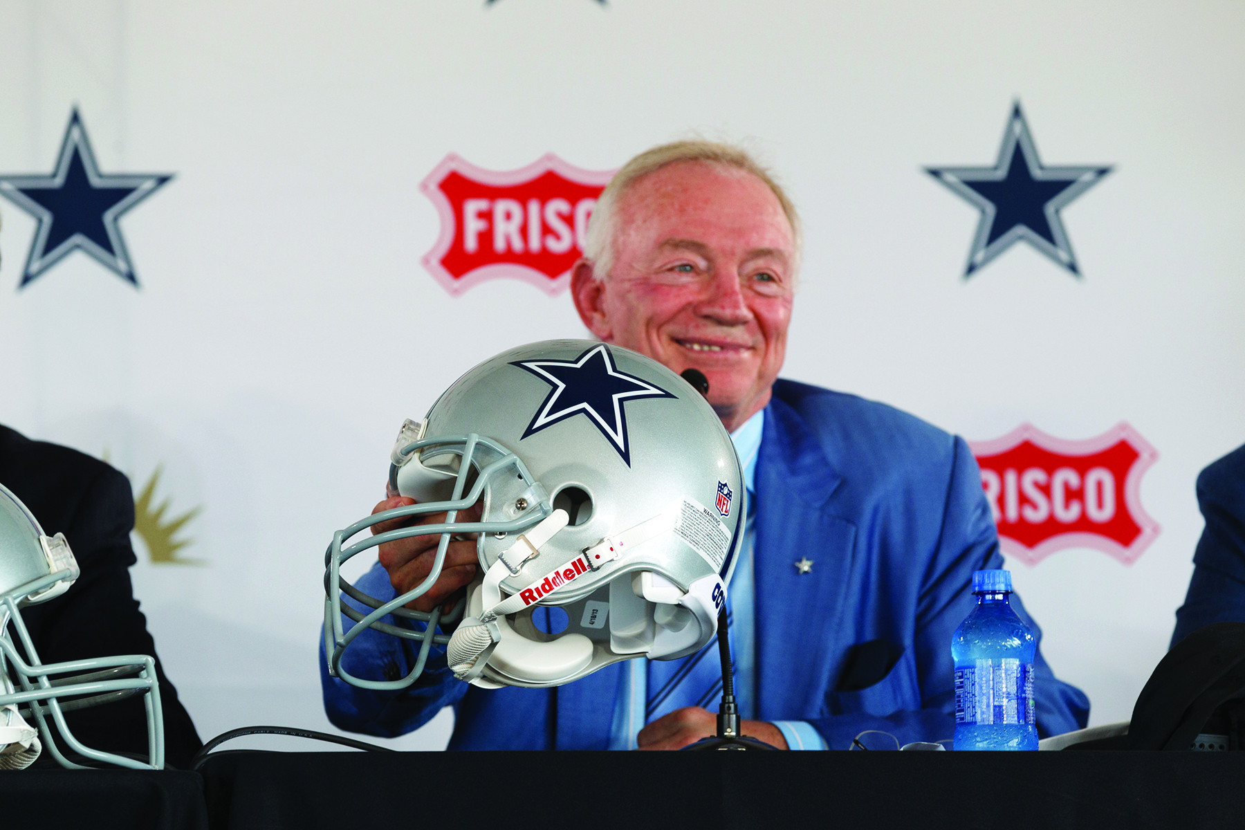Jerry Jones sits at a table for a press conference holding out a silver Dallas Cowboys helmet.