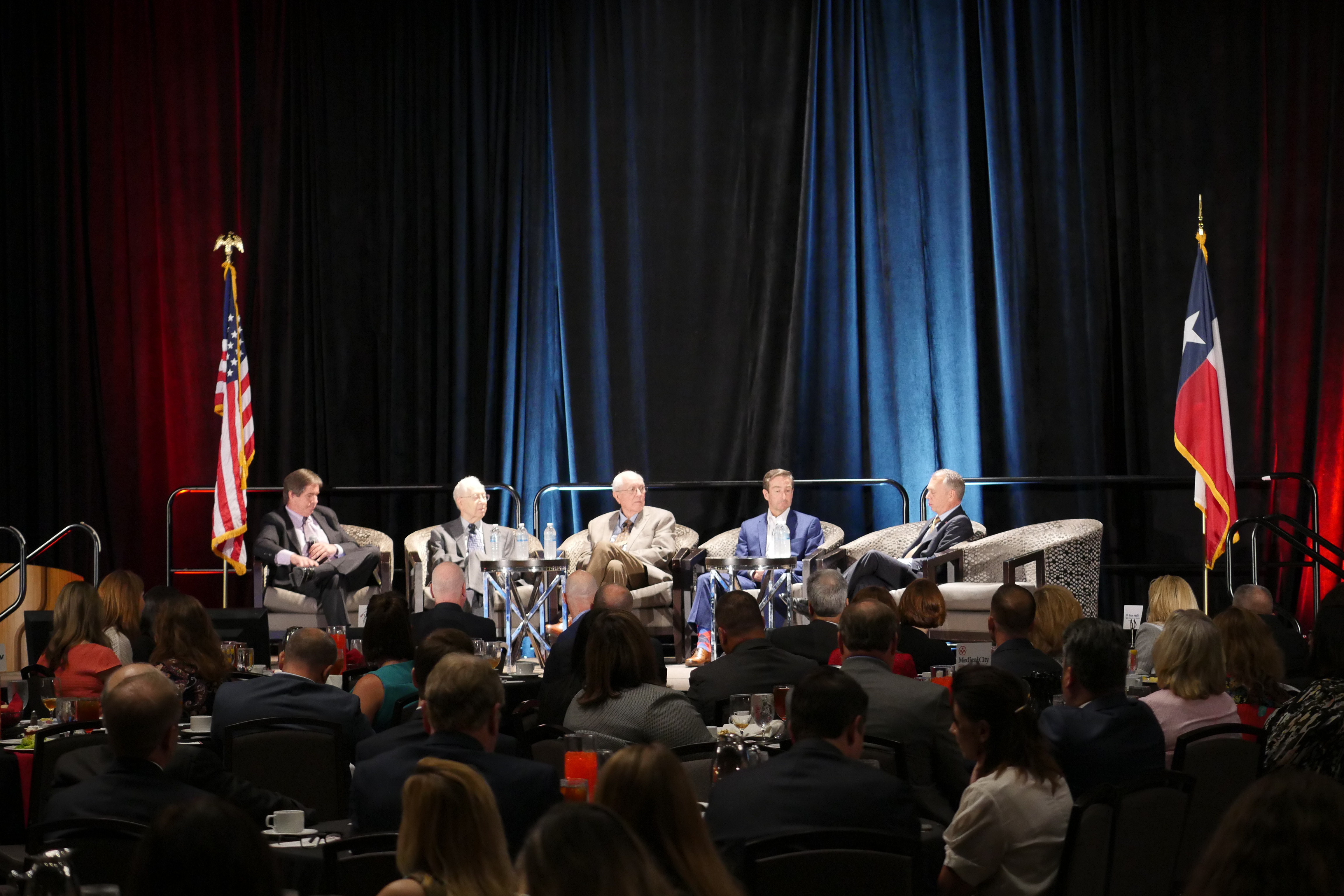 Five men sit in individual chairs on stage at a talk. The crowd is in the foreground.