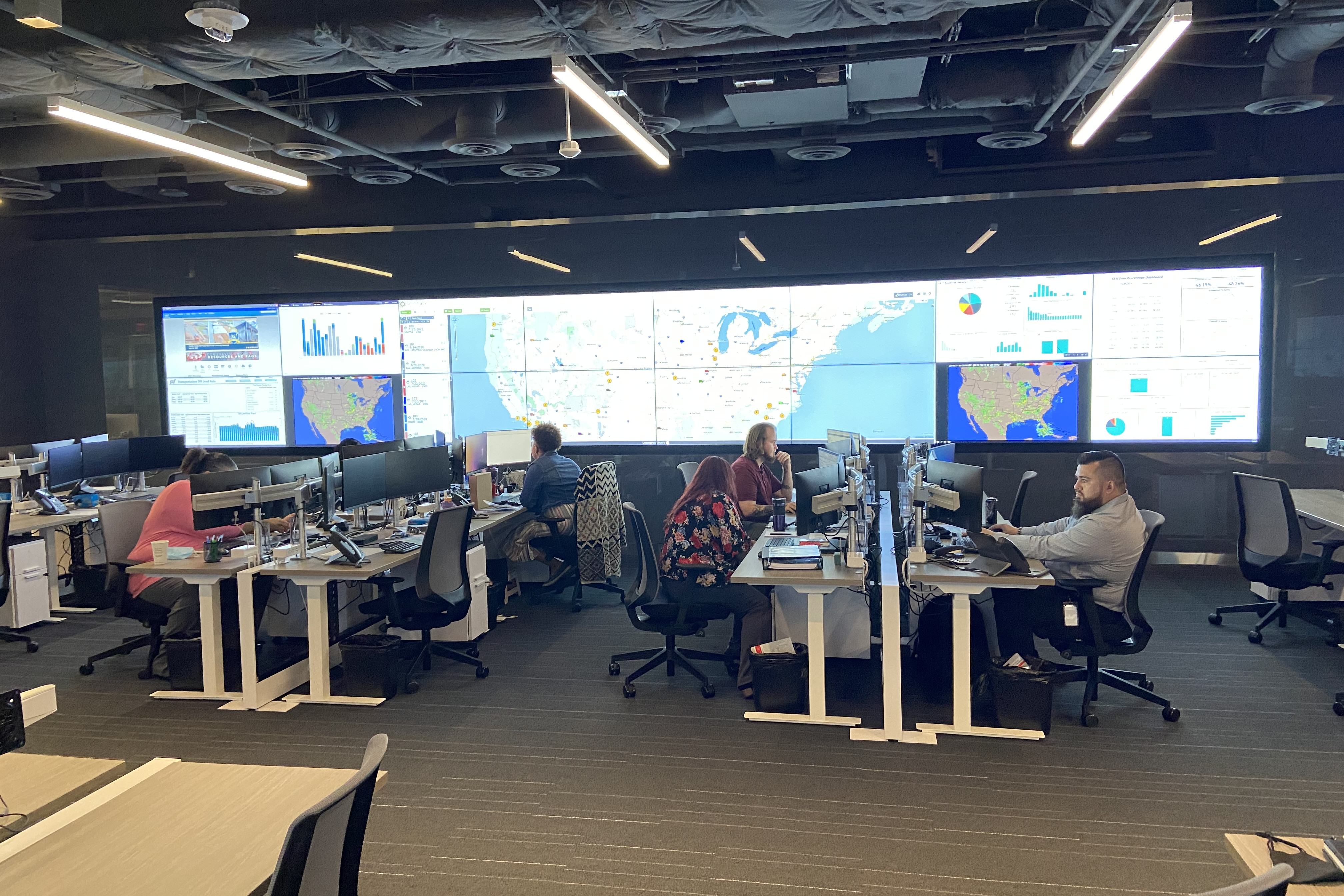 Inside an office, there are rows of employees sitting as desks looking at their computers. There are 16 tv screens at the front with various kinds of information on them