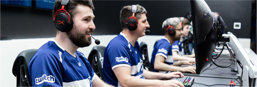 Four men wearing matching blue shirts play video games on their individual computers. They wear matching black and red headsets.