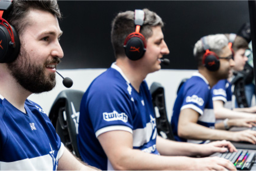 Four men wearing matching blue shirts play video games on their individual computers. They wear matching black and red headsets.