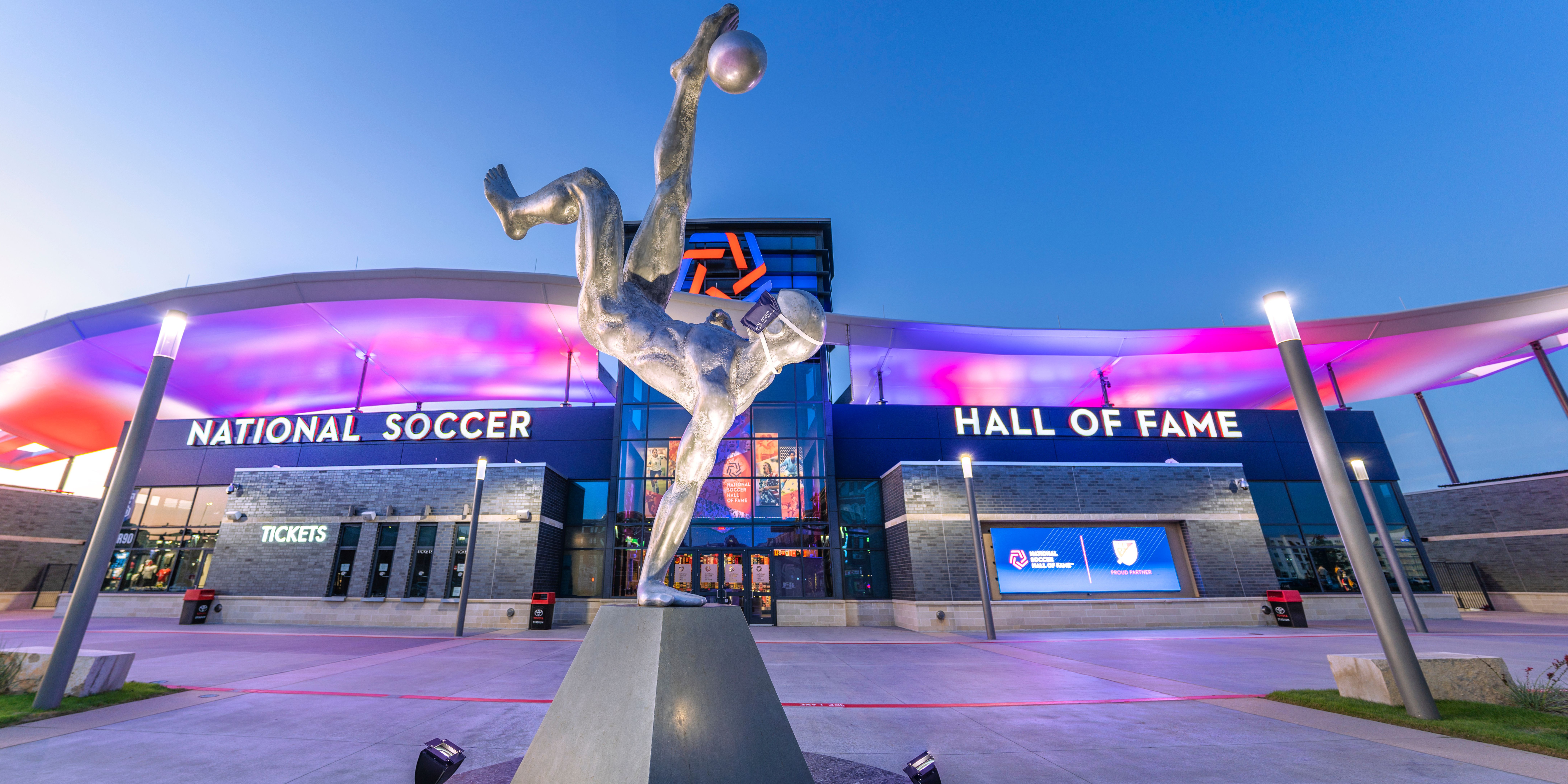 A statue kicking a soccer ball at the front of the National Soccer Hall of Fame. The building is lit up with red and blue lights.